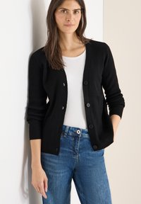 Czarny sweter rozpinany o prążkowanym splocie, z dekoltem w serek, sześcioma guzikami na przodzie i luźnym krojem, zestawiony z białym t-shirtem i niebieskimi dżinsami.