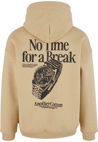 Beige hoodie met een grote zwarte afbeelding van een horloge en tekst: "Geen Tijd voor een Pauze" en "AnotherCotton" in vette letters.
