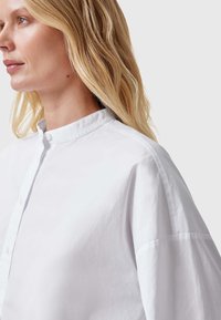 Weißes Baumwollhemd mit Stehkragen, lockerer Passform, Seitennahtdetails und Knopfverschluss vorne. Glatte Textur und minimalistisches Design.
