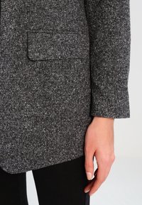 Grå blazer i strukturerad tyg med ett subtilt mönster; har en ficka på bröstet och en åtsittande design. Svart tyg under.