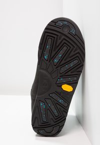 Czarna gumowa podeszwa z bieżnikiem w kształcie zygzaka z niebieskimi plamkami. Widoczne logo Vibram i UGG, zaokrąglony kształt, teksturowana powierzchnia.