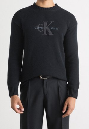 Osoba ubrana w czarny sweter Calvin Klein Jeans z prążkowanymi mankietami oraz czarne spodnie z paskiem, stojąca na tle jednolitego tła.
