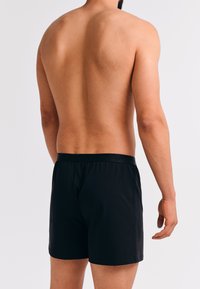 Man met blote bovenlichaam in zwarte bokser shorts, gezien van achteren tegen een effen witte achtergrond.