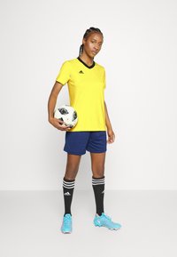 Geltona futbolo marškinėliai su juodais akcentais, tamsiai mėlyni šortai, juodos kelio kojinės su baltais dryžiais ir šviesiai mėlyni sportbačiai, laikantis raštuotą futbolo kamuolį.