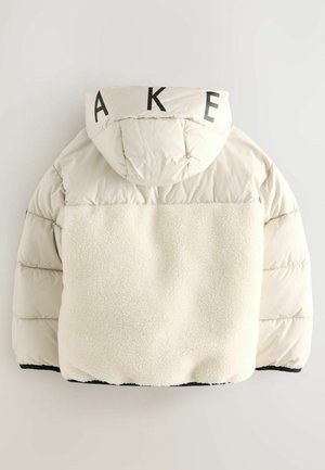 Nafúkaná bunda béžovej farby s textúrovaným krémovým fleece vzadu, so štítkom a veľkými čiernymi písmenami "A K E" na kapucni.