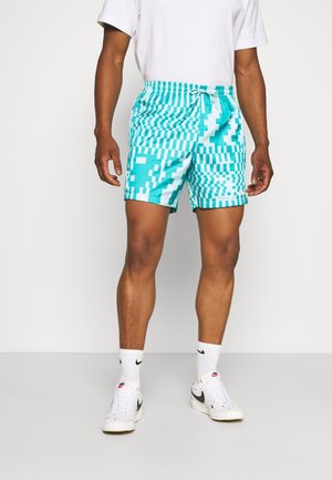 Man met turquoise en wit patroon korte broek, wit T-shirt, witte Nike sokken en witte Nike sneakers, staand op een effen vloer.