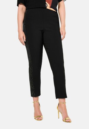 Pantalons noirs ajustés et de longueur cheville, avec des accents métalliques dorés sur les côtés. Portés avec des talons dorés à lanières.