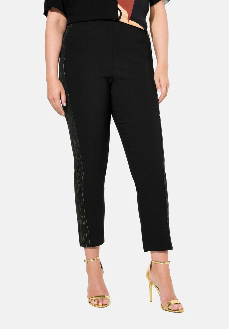 Pantalons noirs ajustés et de longueur cheville, avec des accents métalliques dorés sur les côtés. Portés avec des talons dorés à lanières.