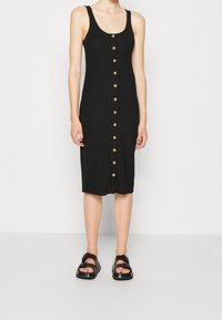 Czarna sukienka z prążkowanej tkaniny, o dopasowanym kroju i długości do połowy łydki, z siedmioma drewnianymi guzikami z przodu; w komplecie z czarnymi sandałami.