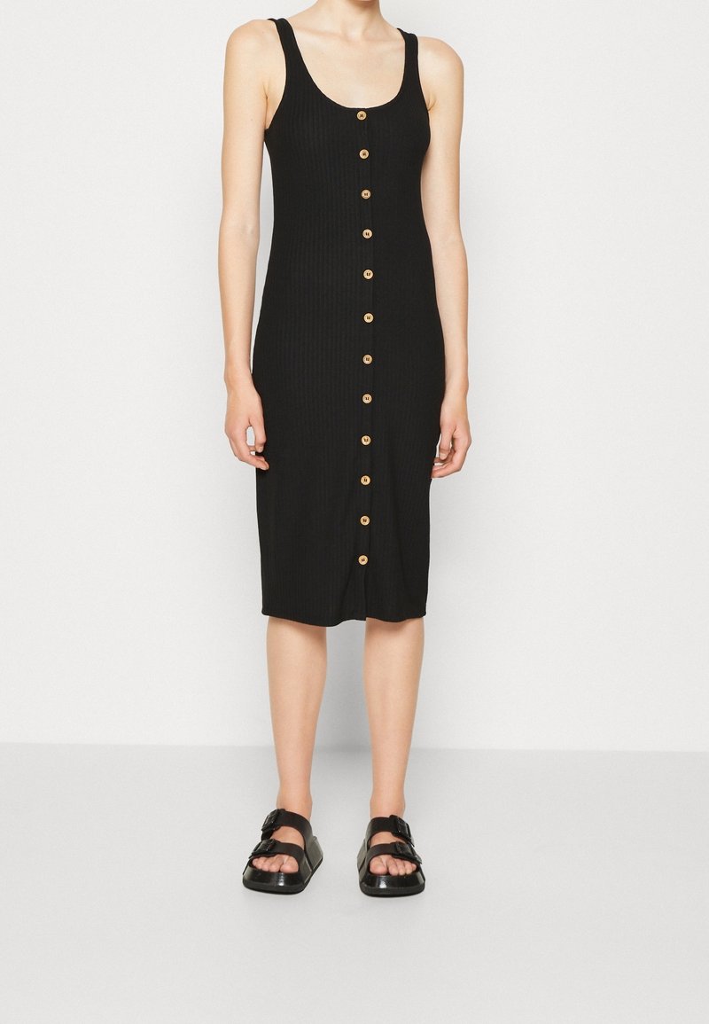 Czarna sukienka z prążkowanej tkaniny, o dopasowanym kroju i długości do połowy łydki, z siedmioma drewnianymi guzikami z przodu; w komplecie z czarnymi sandałami.