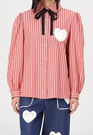Blouse à rayures verticales rouges et roses avec nœud papillon noir, boutons en perle et patch en forme de cœur blanc sur la poche de poitrine ; associée à un pantalon bleu marine avec des patchs en forme de cœur blanc.