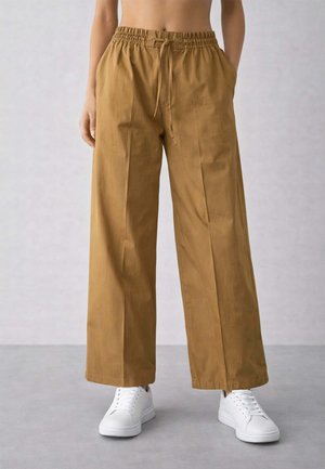 Persona che indossa pantaloni marroni larghi e morbidi con vita elastica e coulisse, mani nelle tasche e scarpe da ginnastica bianche su pavimento grigio.