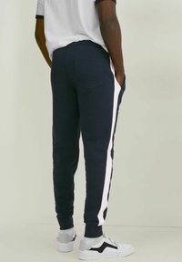 Navy blue collegehousut, joissa on valkoiset sivuhaitarit, joustava vyötärö ja takataskun yksityiskohtia. Pehmeä kangas, jossa on kapeneva lahjetyyli.