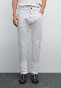 Pantalon en denim blanc avec une coupe slim, doté d'une poche avant et d'une fermeture à bouton supérieur, porté avec des mocassins noirs sur une surface texturée.