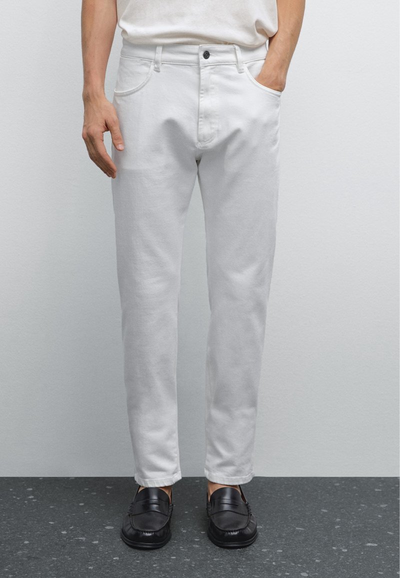 Pantalon en denim blanc avec une coupe slim, doté d'une poche avant et d'une fermeture à bouton supérieur, porté avec des mocassins noirs sur une surface texturée.