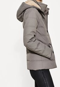 Grå pufferjakke med hætte, der har en beige faux fur-foring, lynlås lukning og lodrette syninger. Sidelommer inkluderet.