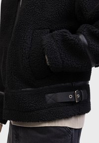Schwarze Fleecejacke aus texturiertem Stoff, mit einem Ledergürtel in der Taille. Lange Ärmel und gerippte Strickbündchen sichtbar.