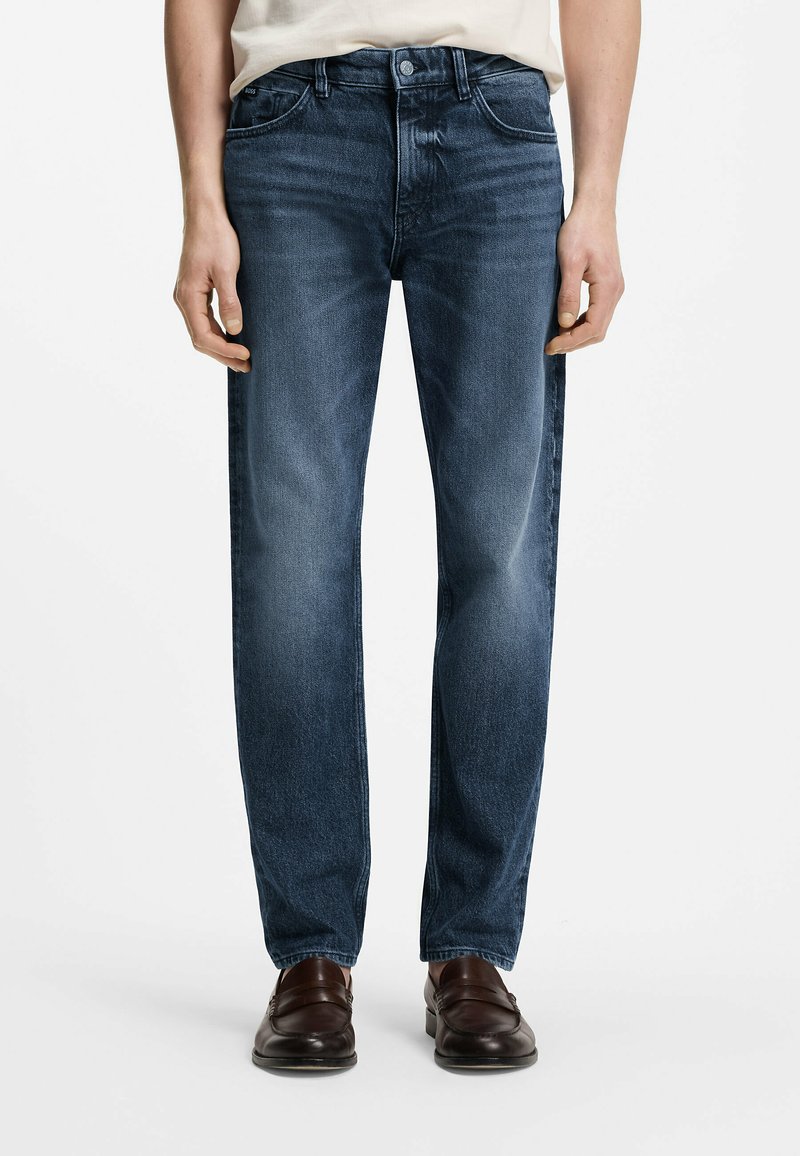 Homme portant un jean bleu délavé droit avec des mocassins en cuir marron, debout devant un fond blanc uni.
