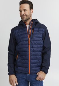 Marineblå puffjakke med quiltet design, oransje glidelås og hette. Har sidelommer og elastiske mansjetter. Laget av syntetiske materialer.