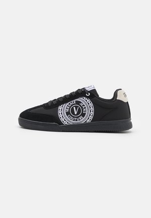 Baskets basses noires avec des lacets noirs, logo circulaire blanc "Versace Jeans Couture" sur le côté, et languette de talon beige clair.