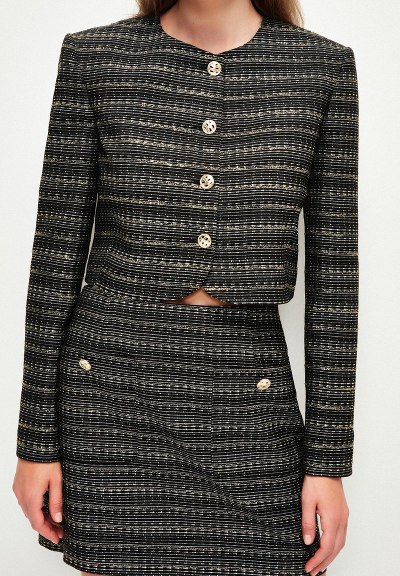 Vrouw in een zwart-wit gestreepte, knoopbare cropped jas en bijpassende, hoge taille rok met goudkleurige knopen.