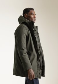 Parka impermeable color verde oliva con capucha, cierre de cremallera y a presión. Cuenta con bolsillos laterales y una textura suave y mate.