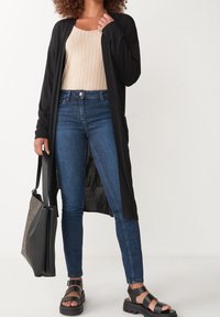 Schwarzer langer Cardigan über einem beigen, gerippten Top, kombiniert mit blauen Skinny-Jeans und schwarzen, offenen Sandalen, hält eine schwarz-graue Tragetasche.