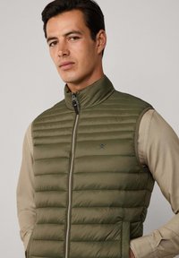 Tummanvihreä, tikattu liivi, jossa on korkea kaulus ja edessä vetoketju. Varustettu kahdella sivutaskulla ja huomaamattomalla logolla rinnassa. Teksturoitu, kevyt kangas.