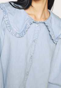 Chemise boutonnée bleu clair en denim, avec un col à volants et des boutons transparents, présentant une texture douce et légèrement texturée.