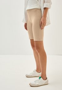 Beige Radshorts aus elastischem Stoff, mit glatter Textur und figurbetontem Design, kombiniert mit weißen Sneakers mit grünen Akzenten.