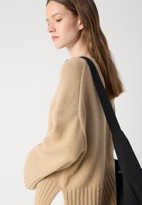 Beige gebreide trui met geribde boorden, relaxte pasvorm en zijsplitten. Gstyled met een zwarte schoudertas met een brede schouderband.