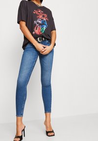 Camiseta gráfica negra con un diseño colorido de los Rolling Stones. Combinada con jeans ajustados azules y sandalias de tacón negras. Collar de plata simple.