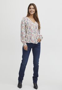 Blusa floreale con maniche lunghe e scollo a V, caratterizzata da uno sfondo bianco e motivi rossi, blu e verdi. Abbinata a jeans blu scuro.