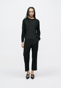 Zwarte hoodie met trekkoorden, gecombineerd met zwarte loszittende broek. Model draagt zwarte puntschoeisel met gespdetails.