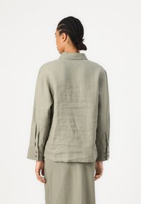Light olive green linen shirt with a collared neck, long sleeves with buttoned cuffs, and a relaxed fit, shown from the back.