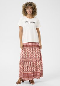 Tricou crem cu text negru "Fă mai mult", asortat cu o fustă maxi roșie cu model. Fusta are un design în straturi și motive florale. Sandale maro.