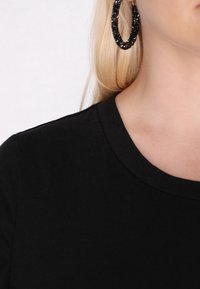 Sort bomulds t-shirt med rund halsudskæring. Glat tekstur synlig, mod en lys baggrund, med store strukturerede sorte hoops øreringe.