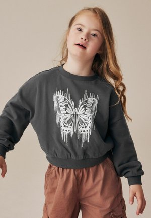 Dunkelgrauer Sweatshirt mit einem weißen Schmetterlingsmotiv, gerippten Bündchen und Saum, kombiniert mit hellbraunen Shorts mit elastischem Bund.