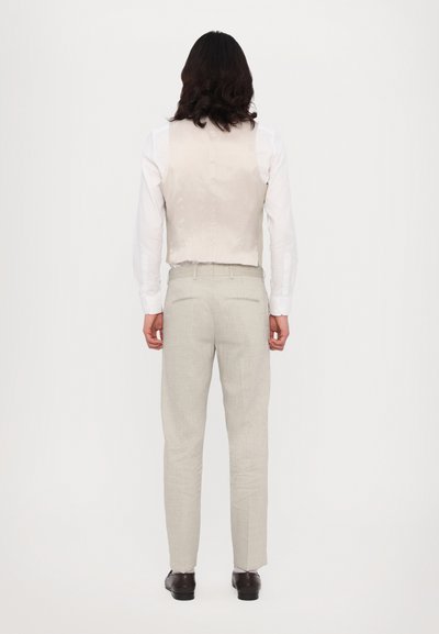 Hellbeige Weste über einem weißen Langarmshirt, kombiniert mit hellgrauen Hosen. Die Schuhe sind aus dunkelbraunem Leder. Gerade Passform.
