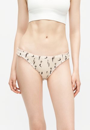 Kobieta w jasnobeżowej bieliźnie typu bikini z małymi czarnymi kwiatowymi i chili drukami oraz białym krótkim topem.