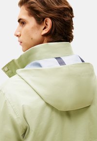 Chaqueta verde claro con capucha, que presenta una textura suave, rayas diagonales en el interior y cierre a presión en el cuello.