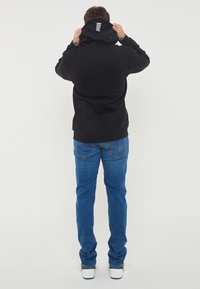 Svart hettegenser med logo på ryggen, paret med blå jeans og hvite joggesko. Stoffet ser mykt og slitesterkt ut.