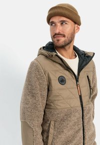 Tan quiltet jakke med fleeceærmer, lynlåslommer og et logo-patch. Sammen med en brun strikket hue med en struktureret overflade.