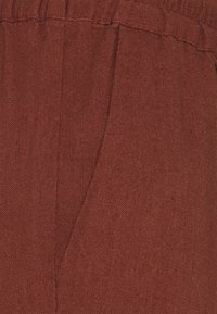 Tissu de couleur rouille avec une surface texturée, présentant une couture latérale et une ceinture élastique visible. Convient pour une tenue décontractée.