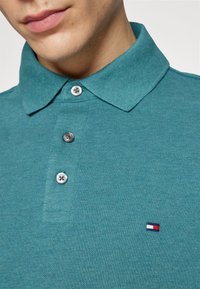 Tyrkysové polo tričko s texturovaným pletením, s knoflíkovým límcem, třemi knoflíky a malým výšivkovým logem v červené, bílé a modré barvě.