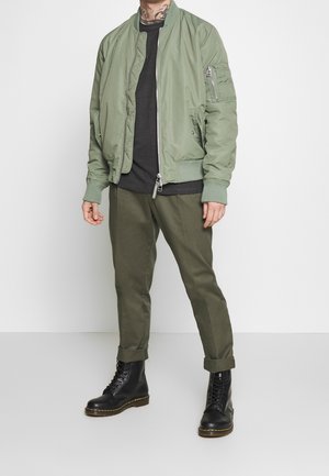 Olivově zelená bomber bunda se zipem vpředu, bočními kapsami a žebrovanými manžetami, spárovaná s tmavě zelenými kalhotami a černými šněrovacími botami.