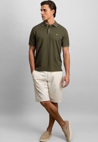 Donkergroene polo met een kraag en een logo, gecombineerd met lichtbeige shorts. Het model staat met één hand in de zak, gekleed in beige instapschoenen.
