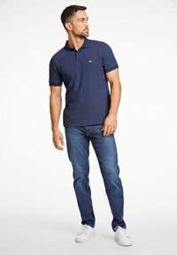 Mörkblå pikétröja med kort ärm, tillverkad av texturerad tyg, kombinerad med blå jeans och vita sneakers. Avslappnad passform, diskret logotypdetalj.