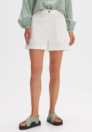 Femme portant un short blanc retroussé avec de grandes poches devant, un chemisier vert à manches longues et des sandales vertes avec des semelles noires.