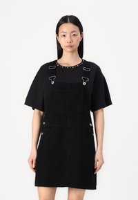 Zwart denim overall jurk met een losse korte mouw top, voorzien van zilveren hardware, zakken en een decoratieve gestikte halslijn.